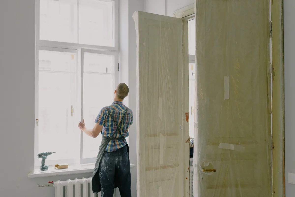 A worker fitting a window from inside a home, a cordless drill resting on the sill